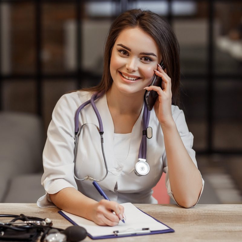 Beautiful young female medical and private doctor is looking at camera and smiling while give a recipe during call